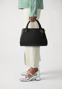 Black textured handbag with logo, gold hardware, and a keychain accent. Worn with silver heeled shoes and pale pant, held in a hand.