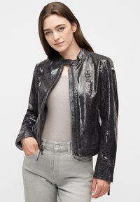 Νεαρή γυναίκα με μακριά καστανά μαλλιά, που φοράει ένα λαμπερό μαύρο μπουφάν με σχέδιο δέρματος φιδιού, μπεζ ριγωτή μπλούζα και ανοιχτόχρωμα γκρι τζιν.