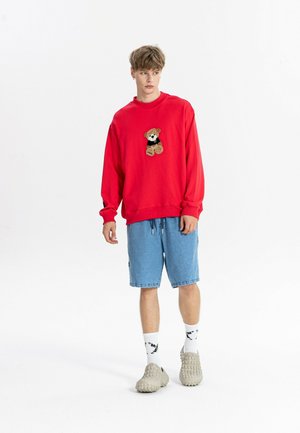 Joven con sudadera roja con un dibujo de osito, pantalones cortos vaqueros azules, calcetines blancos y zapatillas beige texturizadas sin cordones.