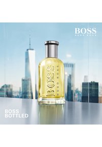 Bouteille en verre transparent avec un capuchon argenté, ornée d'un texte en relief "BOSS HUGO BOSS" et d'un liquide jaune. Mise en scène contre un arrière-plan de skyline urbain.