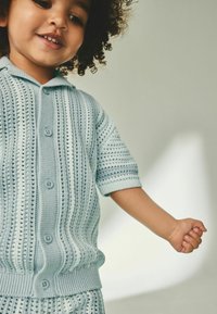 Un pull tricoté bleu clair à manches courtes, avec un motif texturé et une fermeture à boutons, s'accroche au bras d'un enfant.