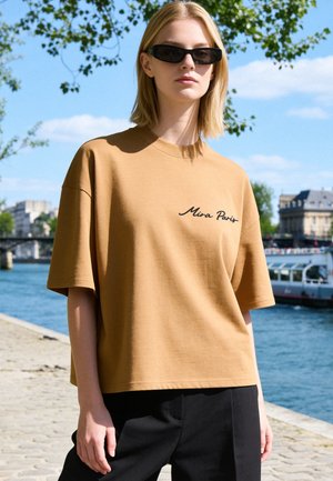 Blonde woman wearing black sunglasses and a brown shirt with "Mira Paris" text stands by a river with buildings and a boat in the background.