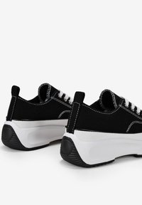 Baskets en toile noires avec des lacets blancs, semelle épaisse blanche, coutures contrastantes et languette arrière en boucle pour un enfilage facile. Semelle extérieure en caoutchouc texturé.