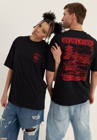 Zwei Personen tragen schwarze T-Shirts mit roten, naturbezogenen Grafiken und Texten und stehen Rücken an Rücken vor einem neutralen Hintergrund.