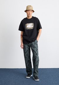 Svart t-skjorte med grafisk trykk, løs passform; grønne mønstrede bukser; beige bøttehatt; svarte sneakers med hvite såler; uformell stil.