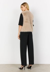 Weste aus Kunstfell in hellbeige, mit einer weichen Textur. Getragen über einem schwarzen Oberteil und weiten schwarzen Hosen, kombiniert mit schwarzen High Heels.