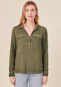 Camisa de manga larga verde oliva con una textura suave, cuello abotonado, dos bolsillos frontales y un dobladillo deshilachado, combinada con un pantalón vaquero azul claro.