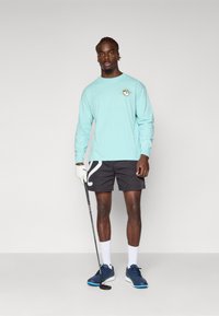 Langermet mintgrønn skjorte med grafikk, svarte shorts med hvitt design, hvite sokker og blå joggesko; holder en golfkølle.