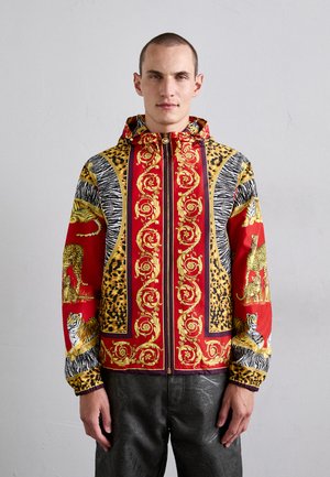 Mand iført en farverig lynlåsjakke med røde, gyldne barokmønstre, dyreprint samt illustrationer af tiger og leopard, der står op ad en ensfarvet væg.