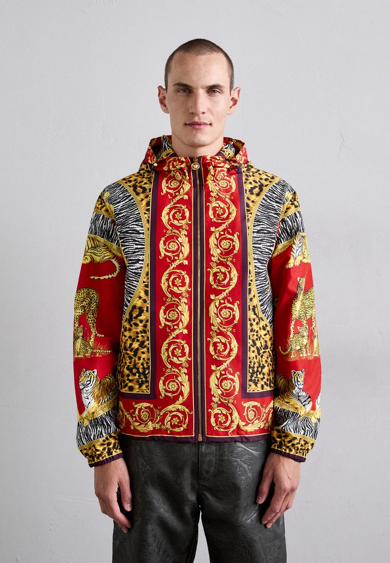 Man som bär en färgglad zip-up jacka med röda, gyllene barockmönster, djurmönster och illustrationer av tiger och leopard, stående mot en enkel vägg.