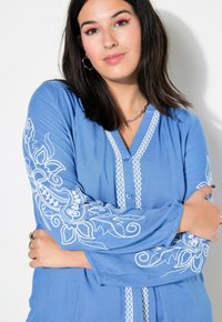 Camicia azzurro chiaro in tessuto morbido con ricami floreali bianchi sui maniche e bottoni decorativi lungo il pannello.