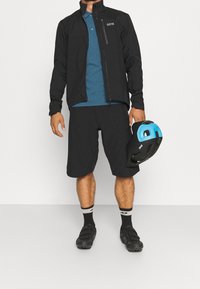 Homme portant une veste noire, une chemise bleue, un short noir, des chaussures de cyclisme noires, et tenant un casque de vélo noir et bleu.