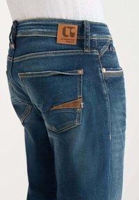 Vaqueros de mezclilla en azul oscuro con una textura suave, que presentan una etiqueta de cuero en la cinturilla y un detalle de bolsillo angular único.