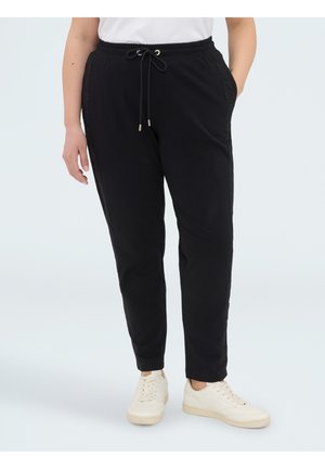 Persoon draagt zwarte joggerbroek met trekkoord en zijzakken en witte sneakers, staand tegen een effen lichte achtergrond.