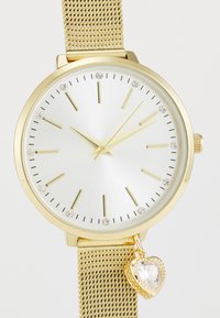 Ρολόι με χρυσή αλυσίδα, στρογγυλή θήκη, λευκό μεταλλικό καντράν, χρυσούς δείκτες ώρων και ένα μενταγιόν σε σχήμα καρδιάς με κρυστάλλινο αξεσουάρ.