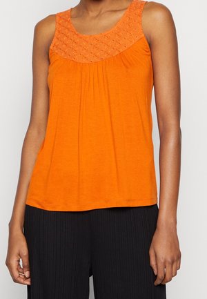 Kvinde iført ærmeløs orange top med blomsterbroderi på bærestykket og sorte plisserede bukser, stående mod hvid baggrund.