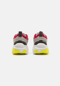 Zapatillas gris y blancas con un diseño robusto, que presentan un interior rojo, acentos negros y una suela amarilla brillante. Marca en el talón.