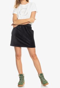 Weißes Baumwoll-T-Shirt mit rundem Logo und Text, kombiniert mit einem schwarzen Cordrock und grünen Winterstiefeln mit fellbesetztem Rand und Schnürsenkeln.