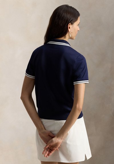 Μπλε σκούρο μπλουζάκι πόλο με άσπρη επένδυση, κοντά μανίκια και εφαρμοστή σχεδίαση. Η πίσω πλευρά έχει ριμπ τελείωμα στο κάτω μέρος και λεία υφή.