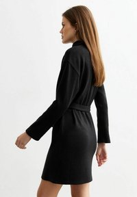 Zwarte wikkeljurk van textuurstof met lange mouwen en een ceintuur in de taille. Heeft een eenvoudig, getailleerd ontwerp en een hem die tot op de knie reikt.