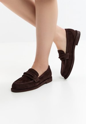 Nahaufnahme von Beinen, die dunkelbraune Wildleder-Loafers mit niedrigem Absatz auf einem weißen Boden tragen.