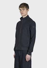 Veste légère noire avec un col montant, fermeture éclair à l'avant, poches latérales et poignets élastiques. Texture lisse et design minimaliste.