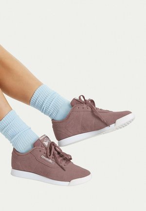 Ben iført lyseblå sokker og lilla Reebok-sneakers med hvide såler, krydsede og løftet mod en ensfarvet hvid baggrund.