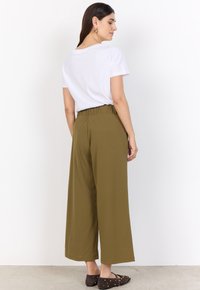 Woman facing sideways wearing a white short-sleeve t-shirt, olive green wide-leg pants, and dark studded flats against a white background.