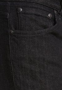 Primer plano de un bolsillo delantero de unos vaqueros de mezclilla negros con remaches de metal y detalles de costura visibles.