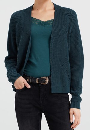 Nainen pukeutuneena petroolinvihreään pitsireunukseen camisoleen, tummanvihreään neulecardiganiin, mustiin housuihin ja mustaan punottuun vyöhön hopeisella soljella.