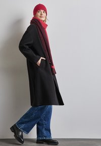 Cappotto di lana nero con tasche, sciarpa rossa lavorata a maglia, cappello coordinato, jeans a gamba larga blu e stivaletti neri con tacco. Sfondo semplice.