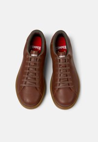 Adidași din piele maro cu o textură netedă, vârf rotund și un design clasic cu șireturi. Prezintă o talpă interioară roșie și o talpă exterioară din cauciuc maro.