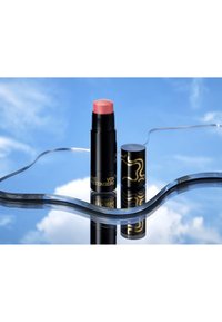 Glanzende zwarte blushstick met goudkleurig ontwerp, dop verwijderd, geplaatst op een reflecterend oppervlak waarop een blauwe lucht en wolken te zien zijn.