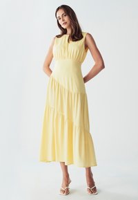 Rumena brezrokavna midi obleka s poševno tiered zasnovo, mehkim materialom in prileganim korpusom. Tokajoča krila z diskretnimi nabrano podrobnostmi.