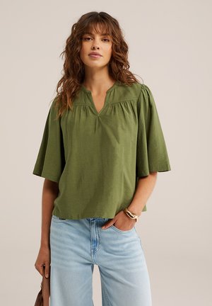 Kobieta z falującymi brązowymi włosami, ubrana w luźną oliwkowozieloną bluzkę z rękawami do łokcia, jasnoniebieskie dżinsy i złotą bransoletkę, trzymająca brązową torebkę.