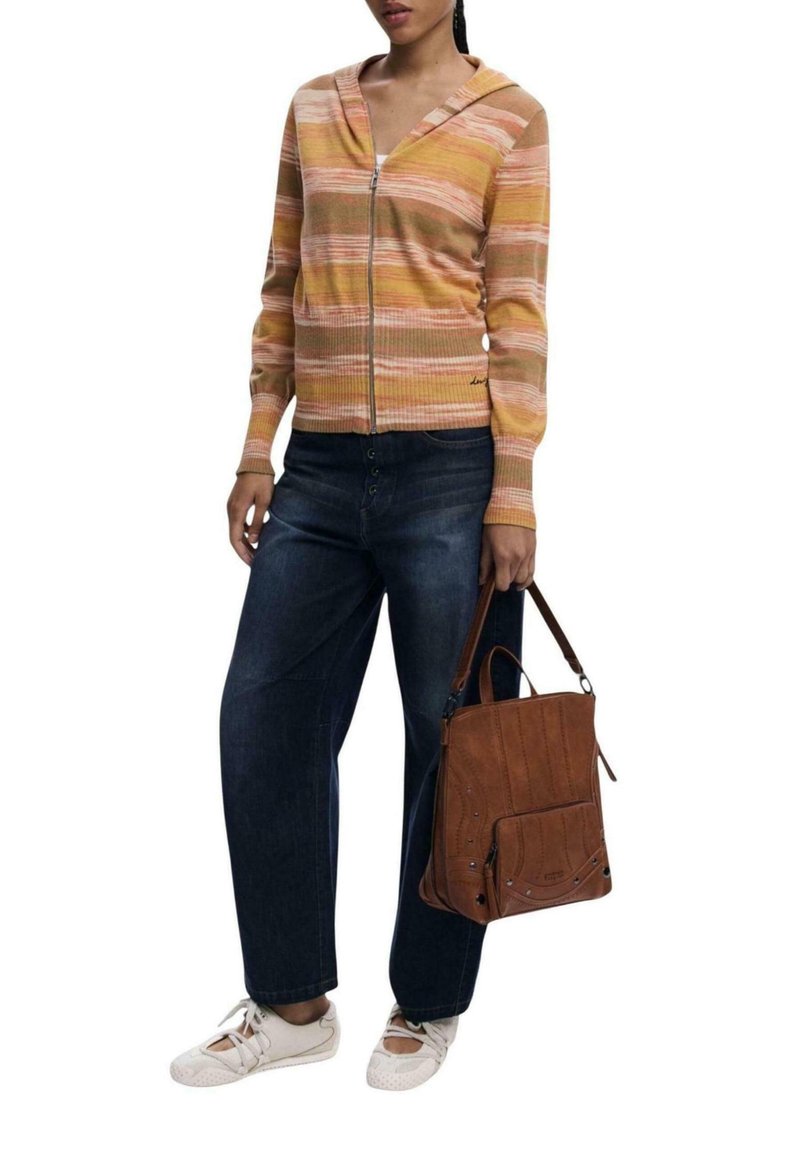 Frau in gestreifter Zip-Jacke, dunklen Jeans, weißen Sneakers und mit einer braunen Lederhandtasche, die sie am Henkel hält.