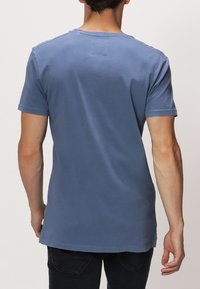 Modrý bavlněný tričko s krátkým rukávem, které má kulatý výstřih, uvolněný střih a viditelné švy podél lemů a výstřihu.