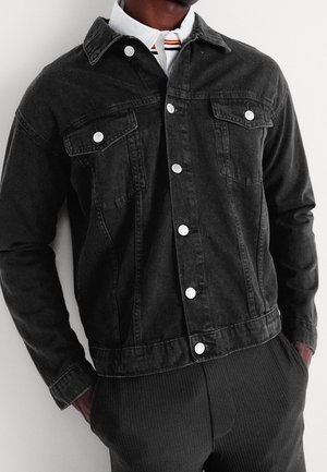 Homme portant une veste en jean noire avec des boutons blancs sur une chemise blanche à col avec des rayures rouges et jaunes.