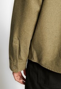 Olijfgroene wolblend jas met een getailleerde snit, voorzien van een knoopmanchet en zijzakdetails, gedragen met zwarte broek.