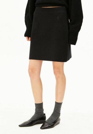 Frau trägt einen schwarzen A-Linien-Rock kombiniert mit grauen Stricksocken und schwarzen spitzen Ballerinas, steht vor einem schlichten weißen Hintergrund.
