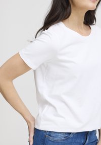 Balti medvilniniai marškinėliai su apvaliu kaklu ir trumpomis rankovėmis, derinami su mėlynais džinsais. Audinys atrodo lygus ir be tekstūros.