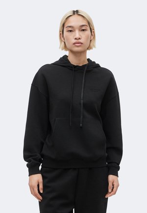 Persoană tânără cu păr blond scurt, purtând un hanorac negru cu buzunar frontal, stând pe un fundal simplu gri deschis.