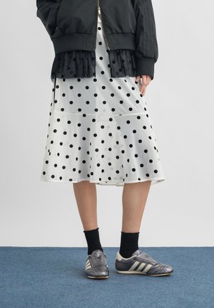 Personne portant une veste noire par-dessus un haut noir à pois, une jupe blanche à pois, des chaussettes noires et des baskets Adidas grises, debout sur un tapis bleu.