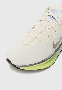 Chaussure de course crème clair avec une tige texturée, un Swoosh Nike gris et une semelle colorée en dégradé passant du jaune au vert.