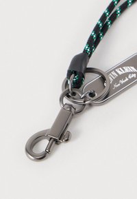 Μπρελόκ με πλεκτό κορδόνι σε μαύρο και πράσινο χρώμα, που διαθέτει μεταλλικό κούμπωμα τύπου αστακού και συνδετικούς κρίκους, με μια ετικέτα που εμφανίζει το όνομα της μάρκας.