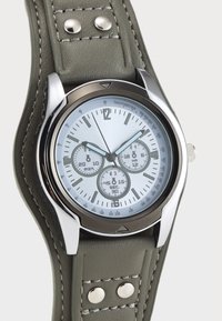 Sølvklokke med lys blå skive, tre underklokker og markante timemarkører. Har en grønn skinnrem med søm og metall detaljer.