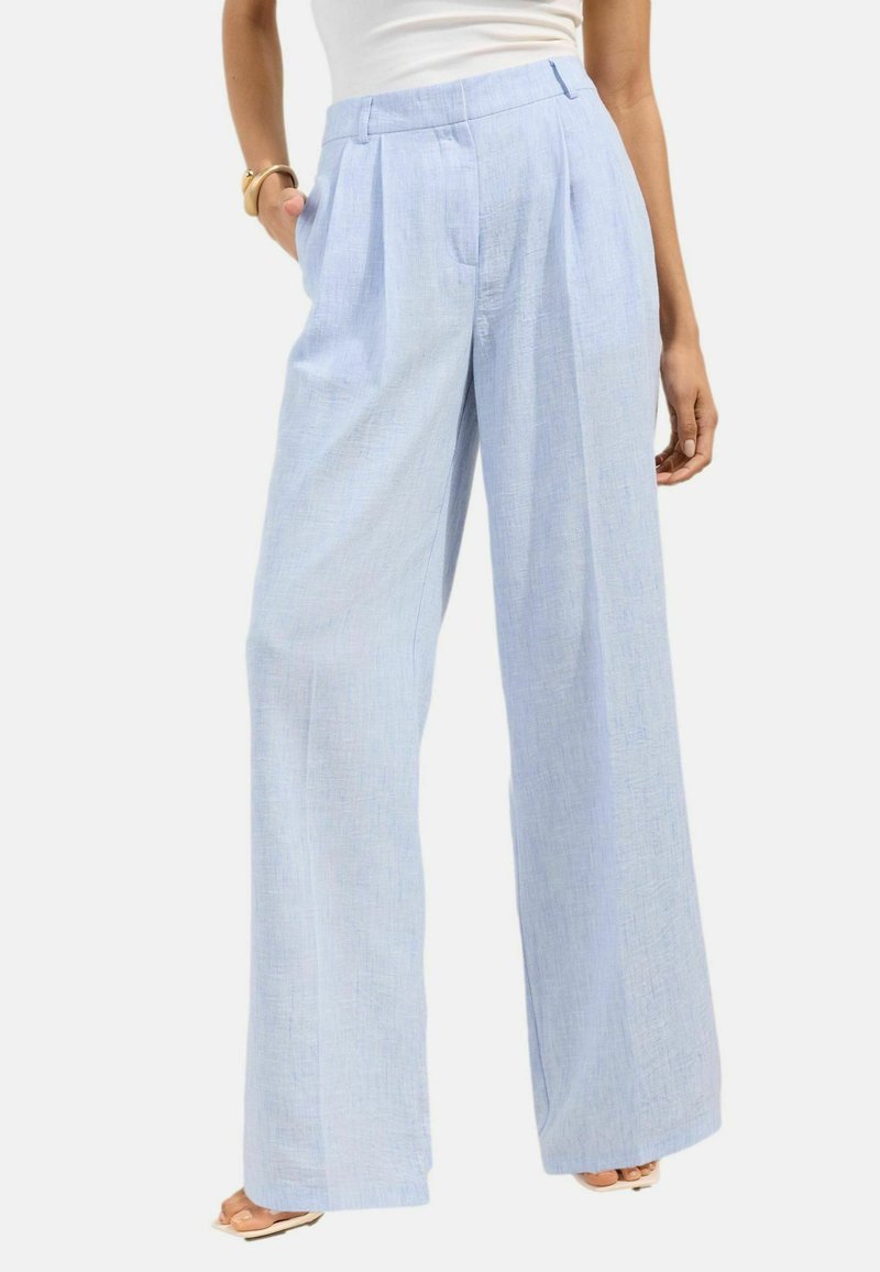 Woman wearing light blue wide-leg trousers with pleats, white tank top, and sandals, standing with one hand in pocket.