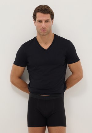 Hombre con camiseta negra de cuello en V y calzoncillos bóxer negros de pie frente a una pared blanca con las manos detrás de la espalda.