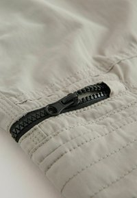 Zbliżenie na czarny zamek błyskawiczny na jasnoszarej tkaninie z widocznym przeszyciem i lekkimi fałdami materiału.