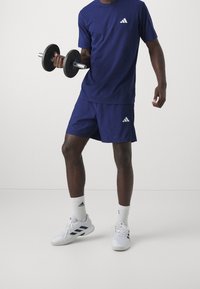 Marinblå sporttröja och shorts med vitt Adidas-logotyp, håller en svart hantel. Vita skor med mörka detaljer, vita strumpor i ankellängd.